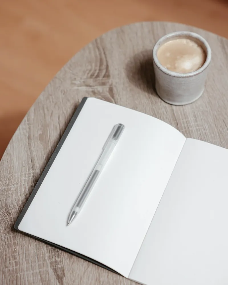 An open notebook with a pen and a cup of coffee on a pale oak desk
